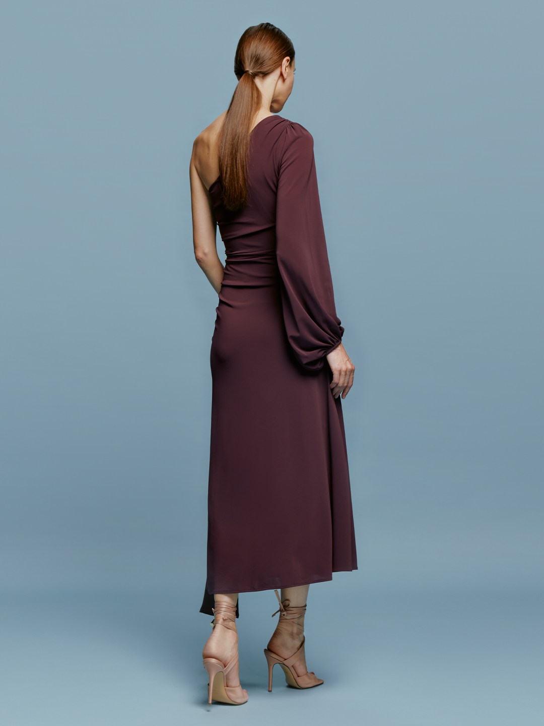 A woman wearing the Mel Dress Burgundy, a one-shoulder dress with ruched detailing and an asymmetrical hem, poses against a plain blue background, capturing a moment of elegance on September 2nd.