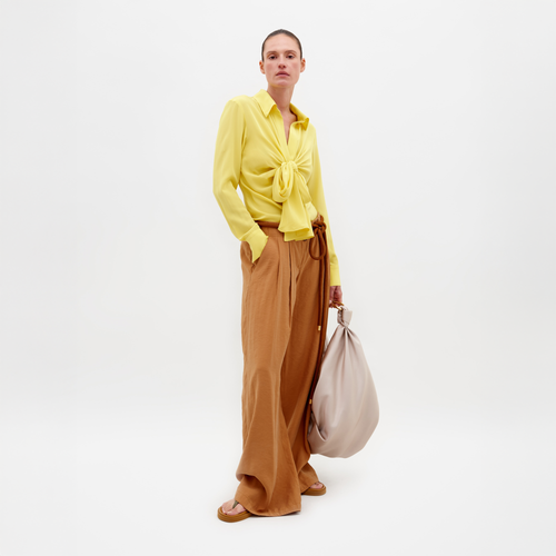 A person stands against a plain background wearing a yellow blouse, the Opal Pant Camel, and sandals, holding a large beige bag. Don't miss the chance to pre-order this outfit by February 15th, 2025.