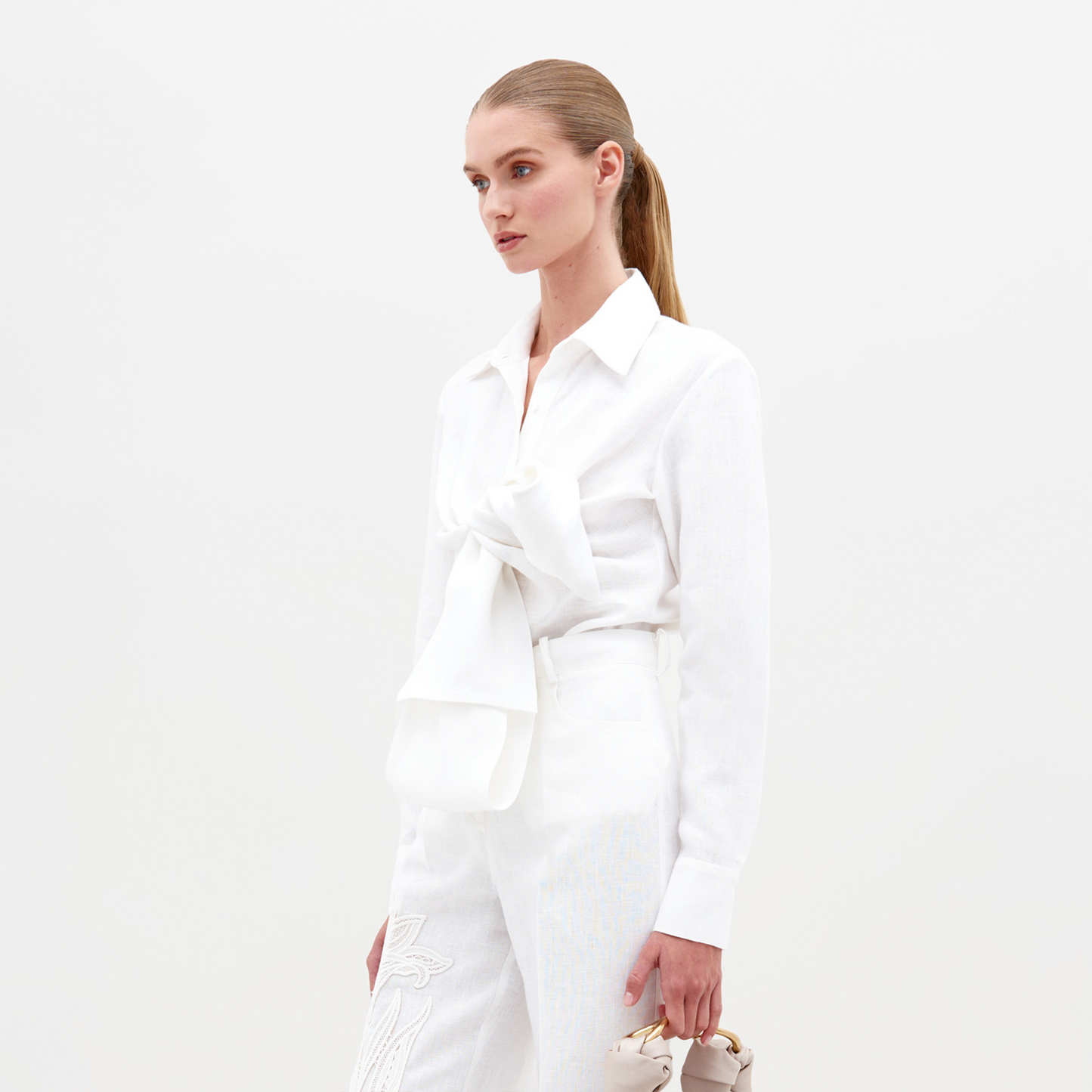 A person with long light brown hair in a ponytail is wearing the Mercury Blouse (Linen) White with a large bow, white pants, and holding beige shoes. Pre-order now to ensure you get this look by the 2025 ship-by date.