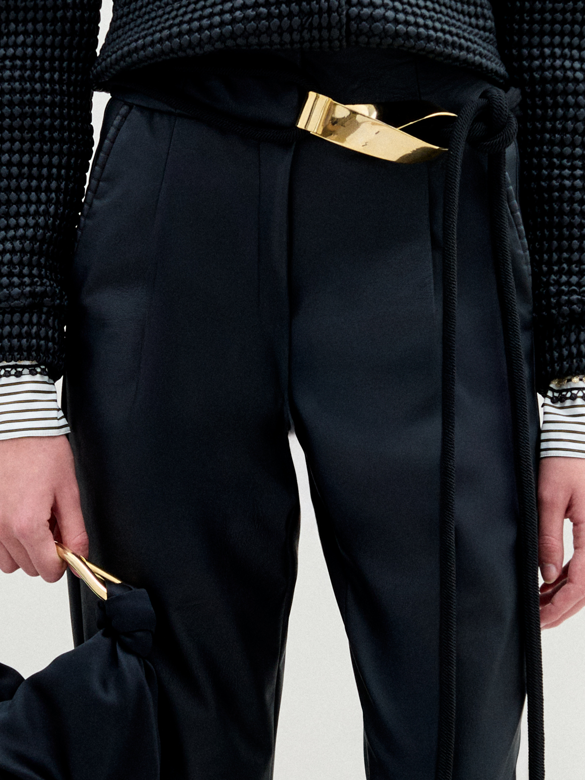 Person stands against a plain white background wearing the Moad Pant, a dark textured jacket, patterned shirt, black boots, and holding a small black handbag.