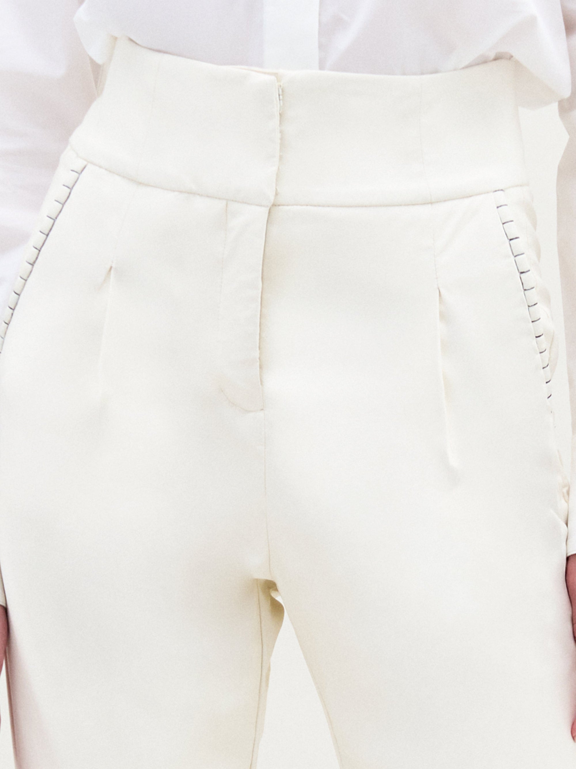 A person stands against a plain light background, wearing a textured white sweater, white collared shirt, the Moad Pant—an Italian fabric high-waisted cropped pant—and white heeled boots.