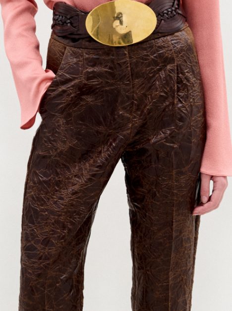 A person stands against a plain background wearing the Moad Pant, a high-waisted cropped style with a straight fitted leg made from Italian fabric, paired with a pink patterned shirt, large gold belt, and black heeled boots.