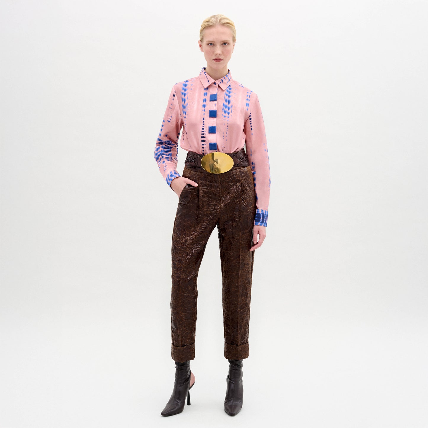 Against a plain backdrop, someone wears the Moad Pant—high-waisted and cropped in Italian fabric with a straight fitted leg—paired with a pink patterned shirt, large gold belt, and black heeled boots.