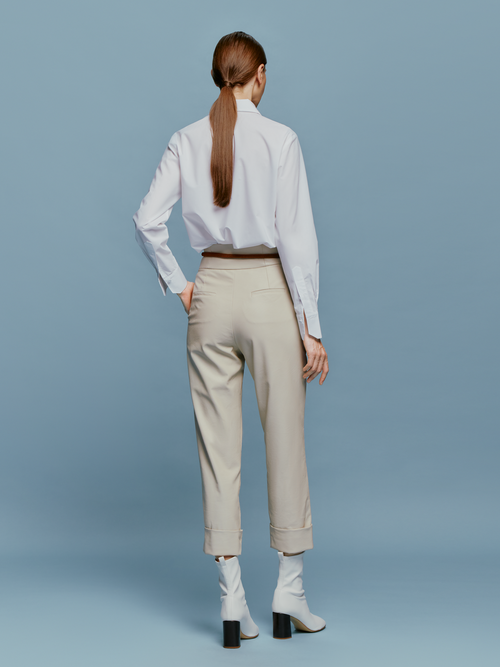 A person stands against a blue background, wearing a white shirt, black tie, Moad Pant Cream high-waisted pants, a brown belt with a keychain dated September 2nd, and white heeled boots.