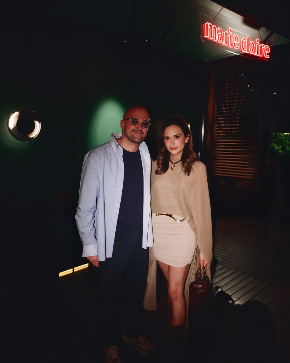Mauricio Espinosa Tcherassi and Sofia Espinosa Tcherassi posing together in a dimly lit venue at night. The woman is wearing a beige long-sleeve top and a matching beige mini skirt with a thin belt, while the man wears a light blue open jacket over a dark shirt and jeans.