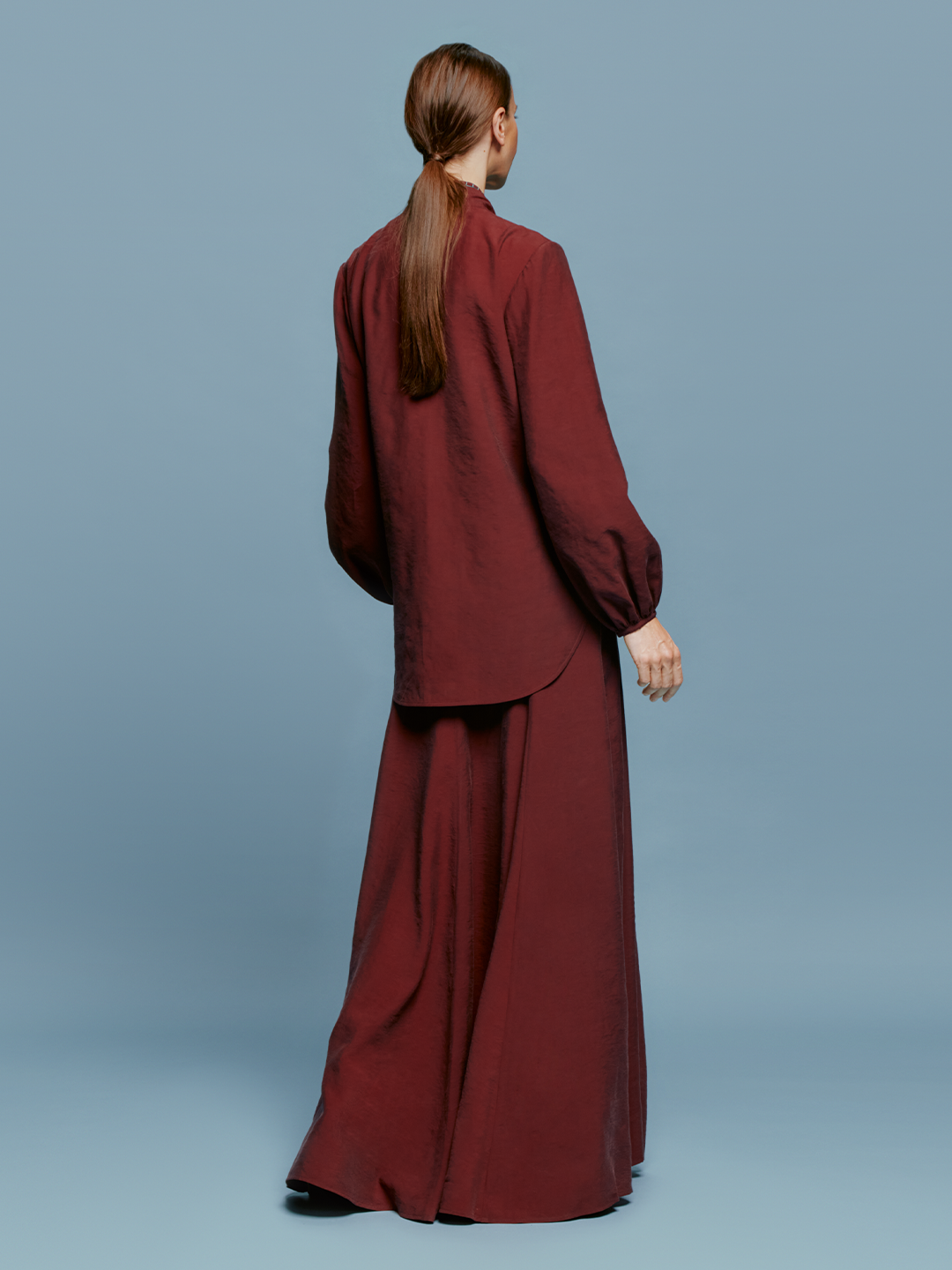 A person in a Modena Skirt Burgundy outfit, consisting of a long-sleeved top with a front tie and a matching long skirt, stands against a plain blue background, evoking memories of summer days on the ship.