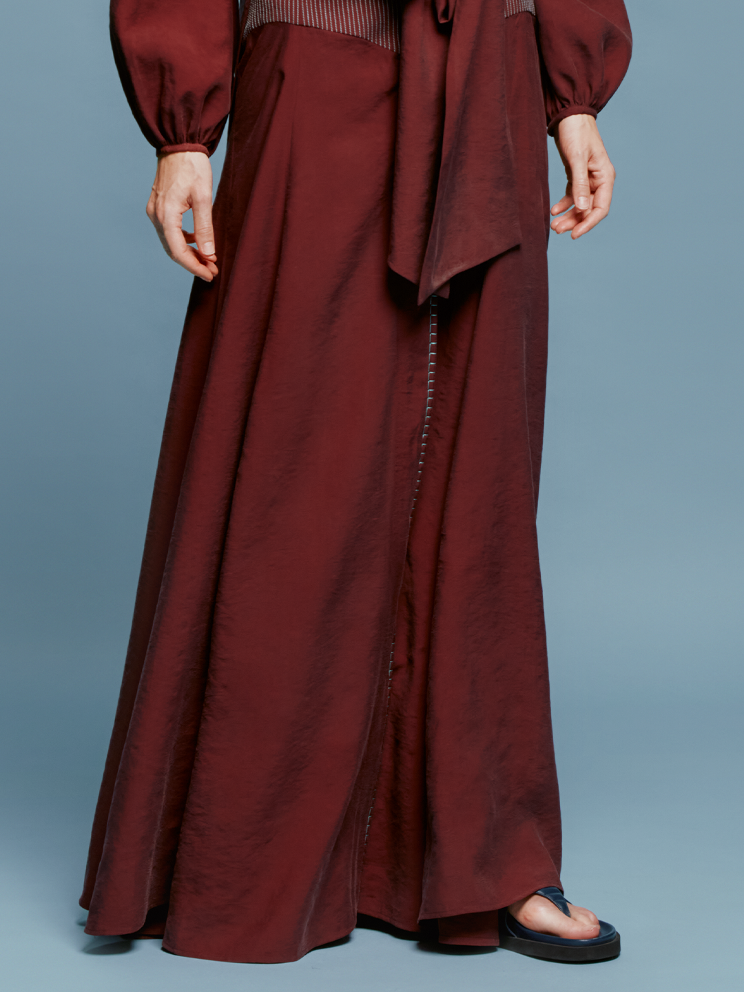 A person in a Modena Skirt Burgundy outfit, consisting of a long-sleeved top with a front tie and a matching long skirt, stands against a plain blue background, evoking memories of summer days on the ship.