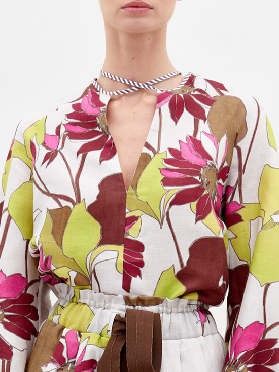 A person stands against a plain background, wearing the Molveno Blouse in Botanical Multifloral, featuring a floral-patterned long-sleeve top with a deep V-neckline and matching high-waisted shorts, cinched at the waist with a brown belt. Pre-order now to ensure it arrives by the February 15th, 2025 ship-by date.