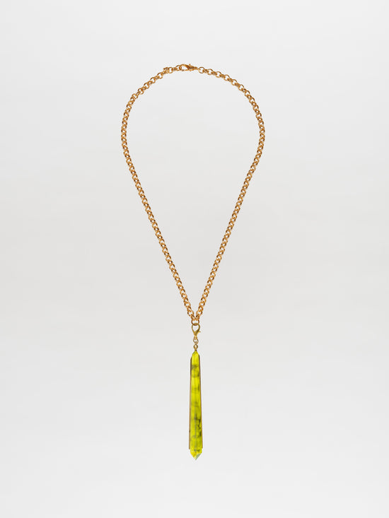 The Montse Necklace Green is a gold-plated brass chain adorned with a long, slender green geometric crystal pendant, elegantly showcased against a plain background.