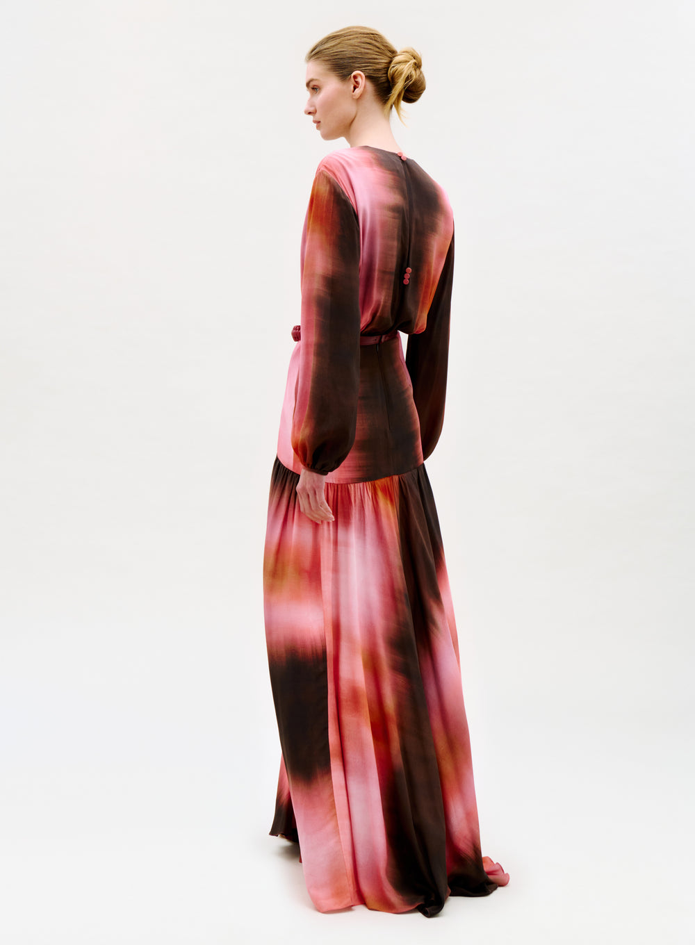 A woman with blonde hair in a neat bun faces away, modeling the Maialen Dress—a long-sleeved, floor-length gown featuring a pink and brown tie-dye pattern—set against a plain white background.
