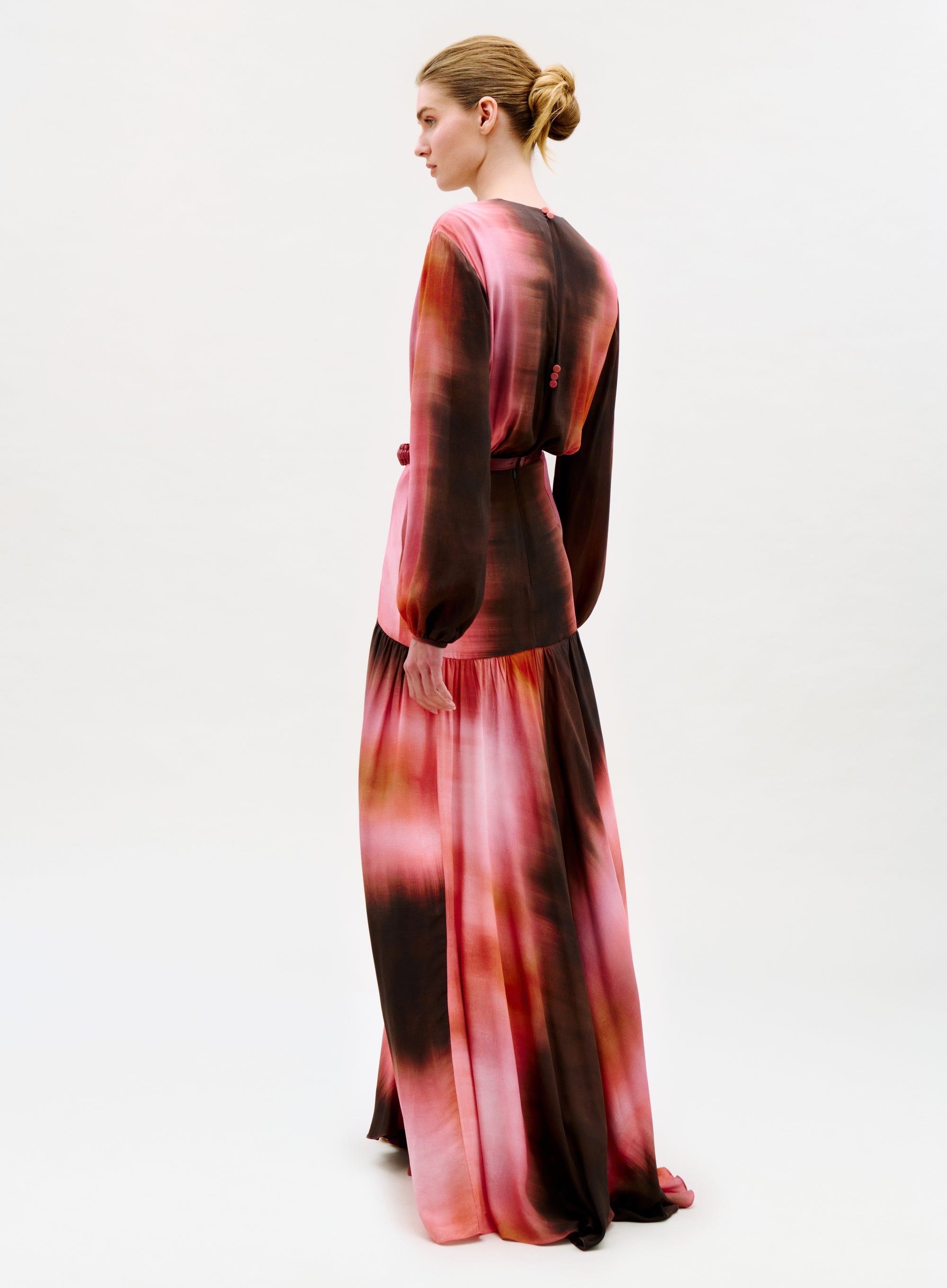 A woman with blonde hair in a neat bun faces away, modeling the Maialen Dress—a long-sleeved, floor-length gown featuring a pink and brown tie-dye pattern—set against a plain white background.