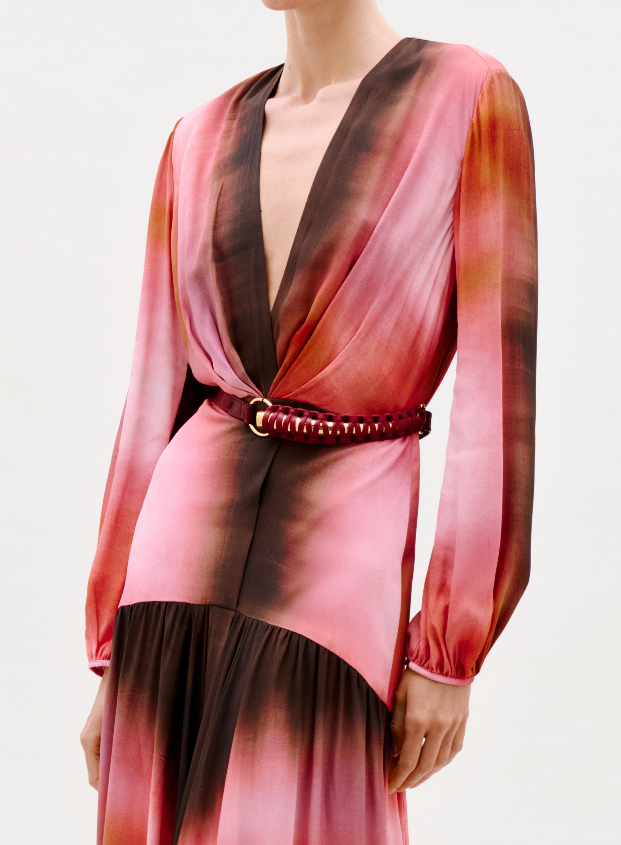 A person wears the Maialen Dress—a long-sleeved, deep V-neck, pink and brown gradient dress with a gathered waist and braided red belt with gold accents. The photo is cropped at the shoulders and mid-thigh.