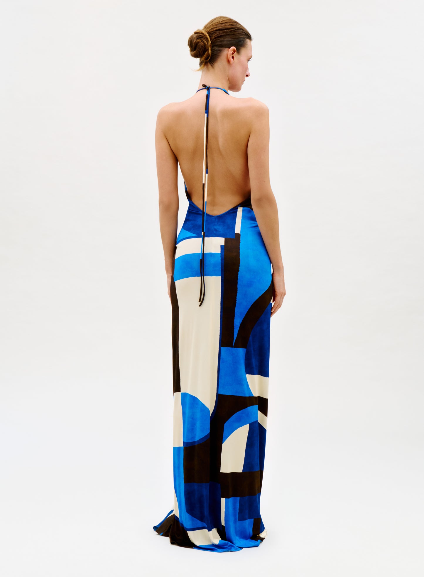 A woman faces away, wearing the Malek Dress—a backless, halter-neck style featuring a bold geometric pattern in blue, black, and white. Her hair is styled in a neat bun against a plain white background.