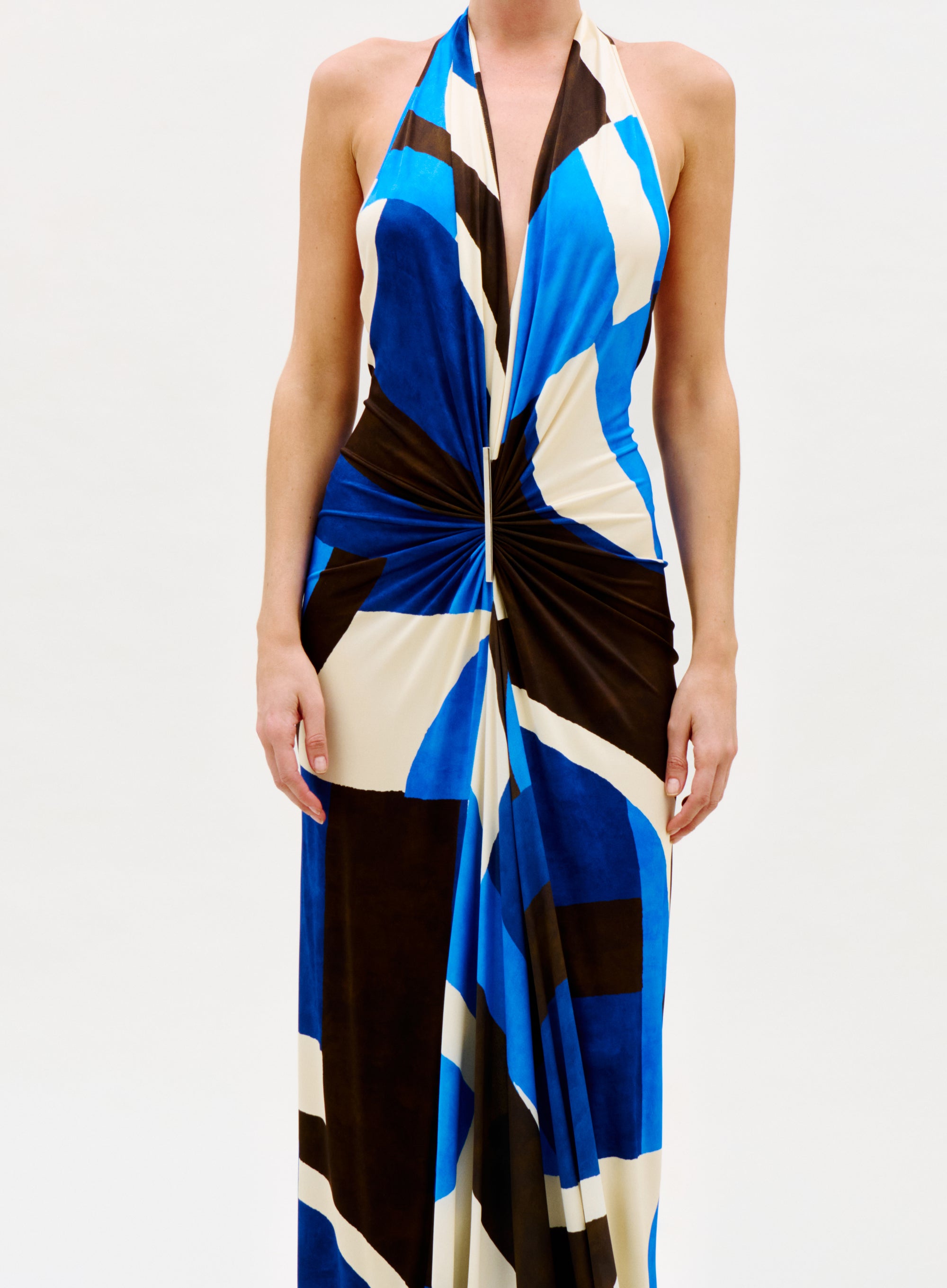 A woman wears the Malek Dress—a sleeveless, halter-neck style with a deep V-neckline, twist detail at the waist, and a bold blue, black, and beige geometric pattern—set against a plain white background.