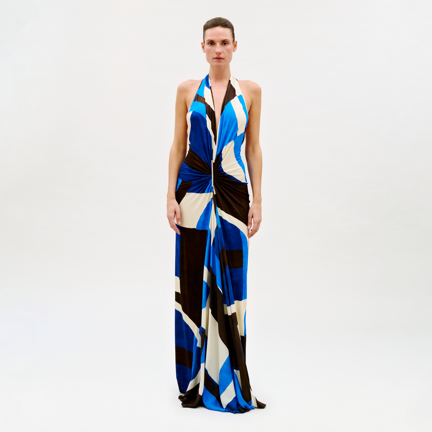 A person poses against a plain light background wearing the Malek Dress, a long halter-neck style with a bold blue, black, white, and beige geometric pattern and a knotted waist detail.