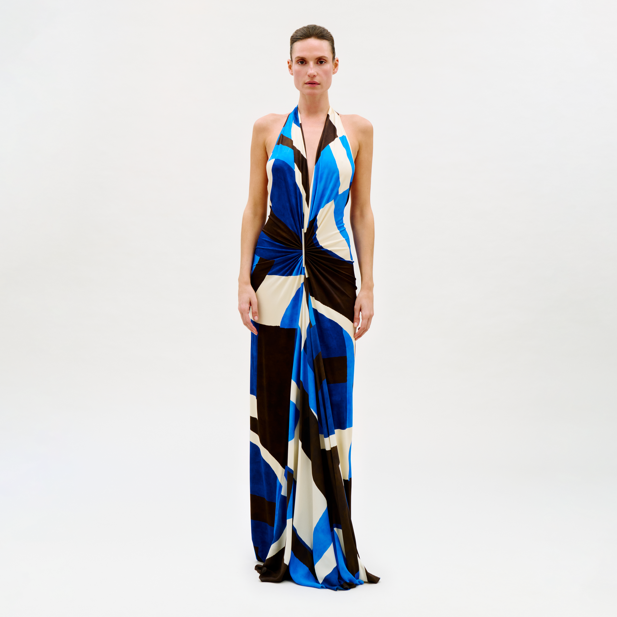 A person poses against a plain light background wearing the Malek Dress, a long halter-neck style with a bold blue, black, white, and beige geometric pattern and a knotted waist detail.