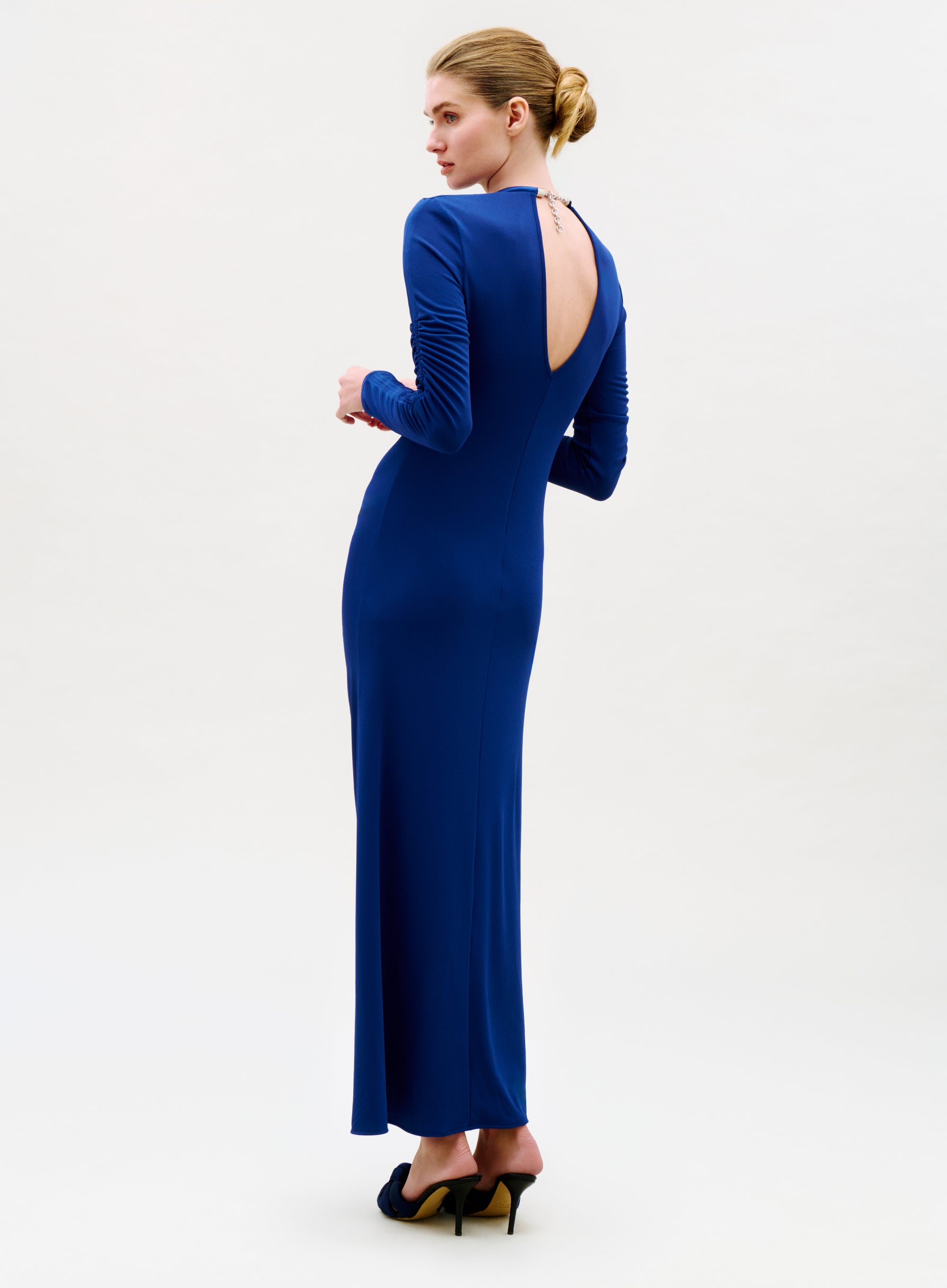 Woman in a long, fitted Mara Dress with long sleeves and an open back, shown in profile wearing high heels and her blonde hair styled in a low bun.