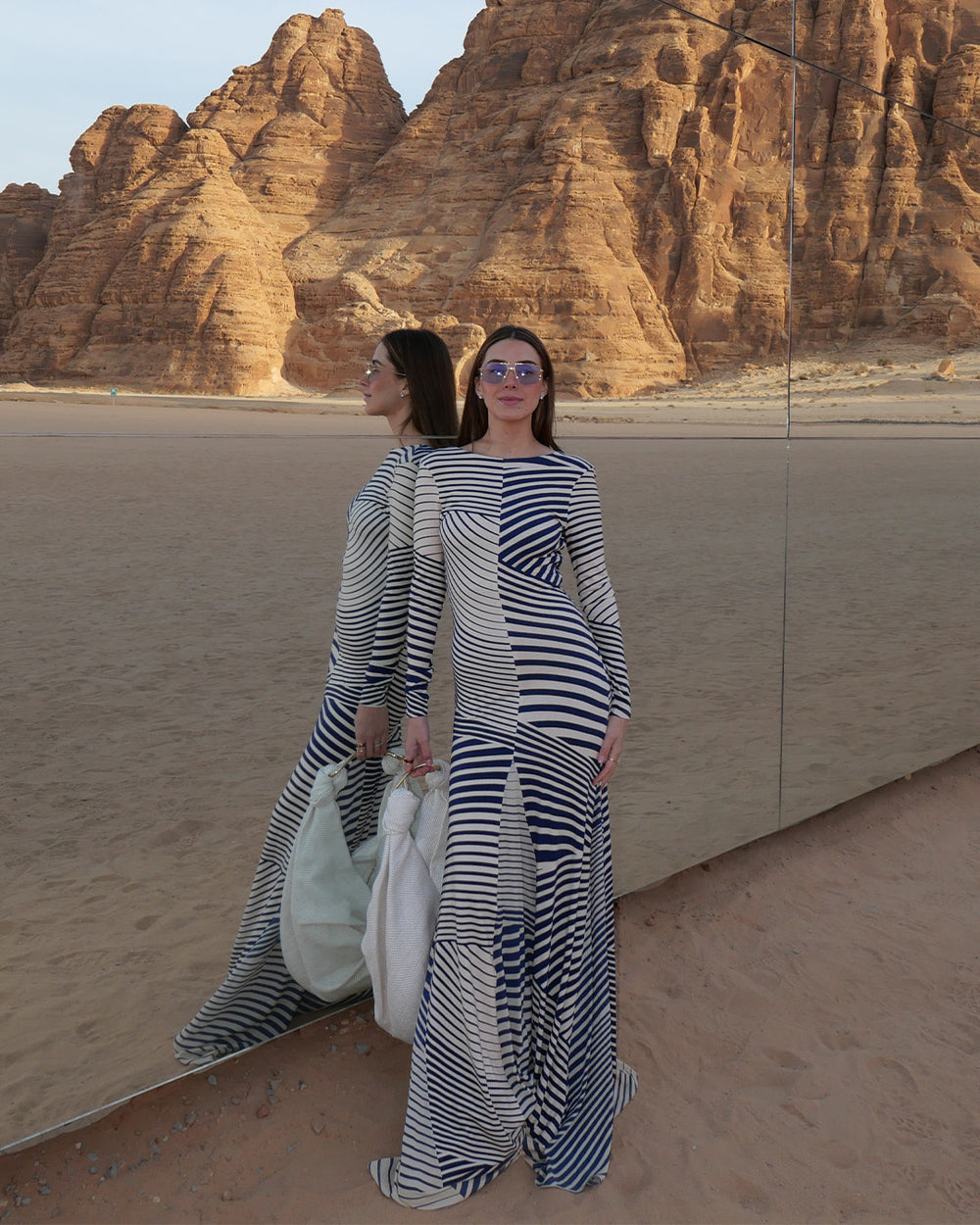 Woman in a striped dress standing in front of a large mirror in a desert setting with rock formations.