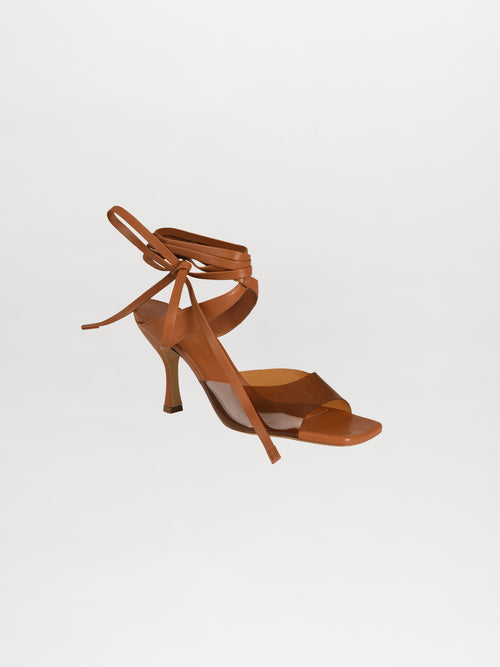 The Marlene Heels feature luxurious Italian calf leather in brown, with high heels, ankle wrap straps, and a PVC transparent strap, set against a plain white background.