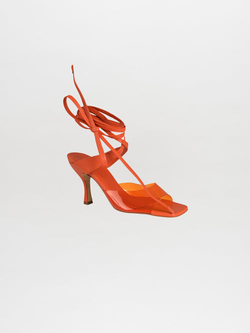 The Marlene Heels Orange feature a bold orange strappy sandal crafted from luxurious Italian calf leather, complete with a mid-heel and sophisticated ankle tie design, elegantly visible from the side.