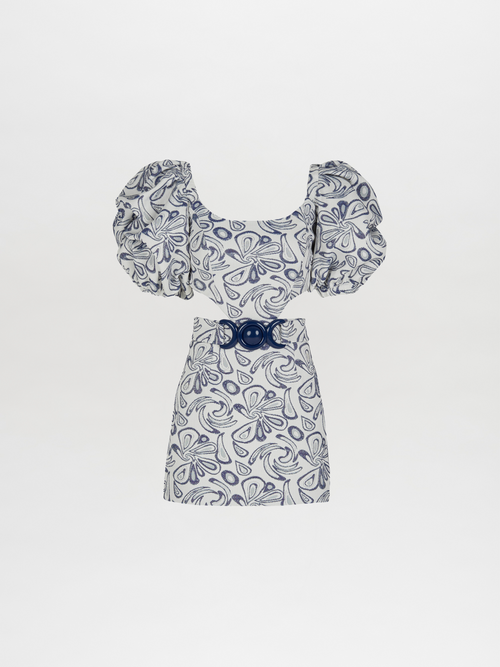 The Marlin Dress Navy Swirls from the Spring 2023 collection is a short white dress adorned with blue abstract patterns, featuring puffed sleeves and a wide navy belt with a large circular buckle.