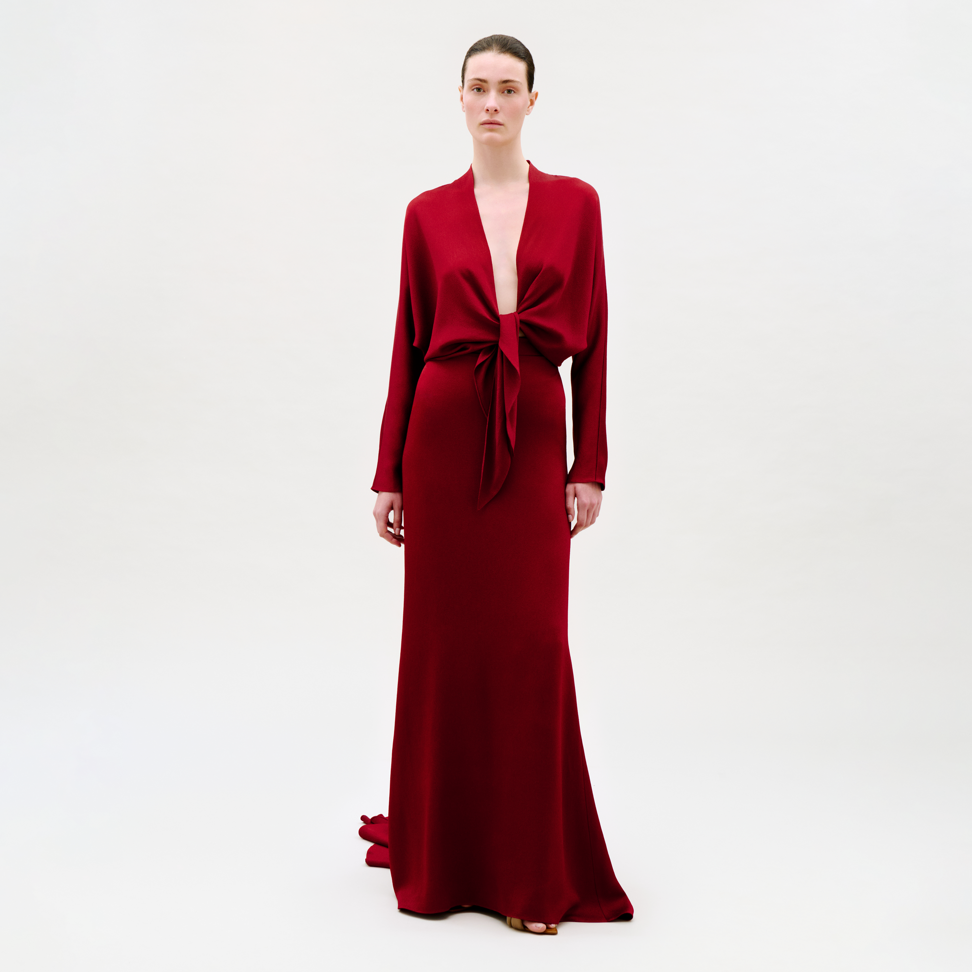 A person stands against a plain background, wearing the Marlon Skirt—a floor-length, deep red skirt styled with a front tie at the waist. The outfit features long sleeves and a plunging neckline for an elegant look.