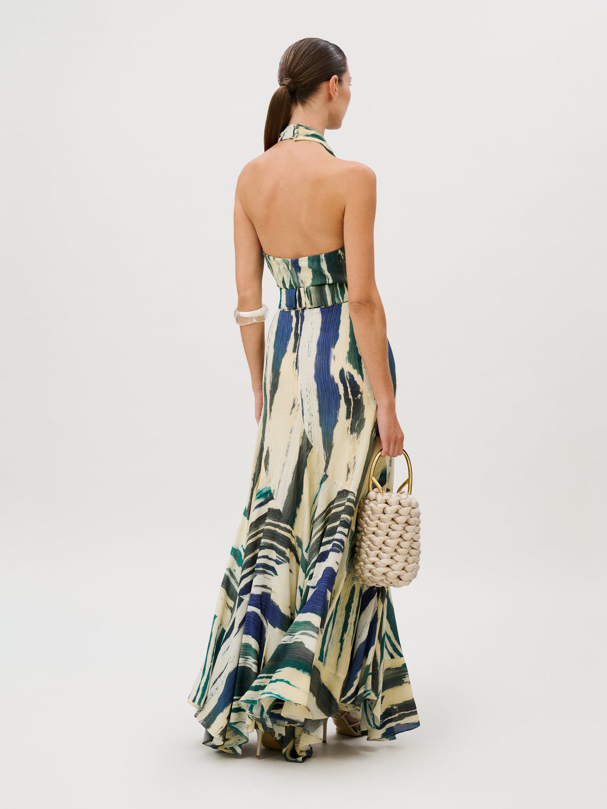 Back view of a person wearing the Marwa Dress, a floor-length crepe de viscose maxi dress with a blue, green, and cream abstract brushstroke print, holding a woven handbag with a gold handle.