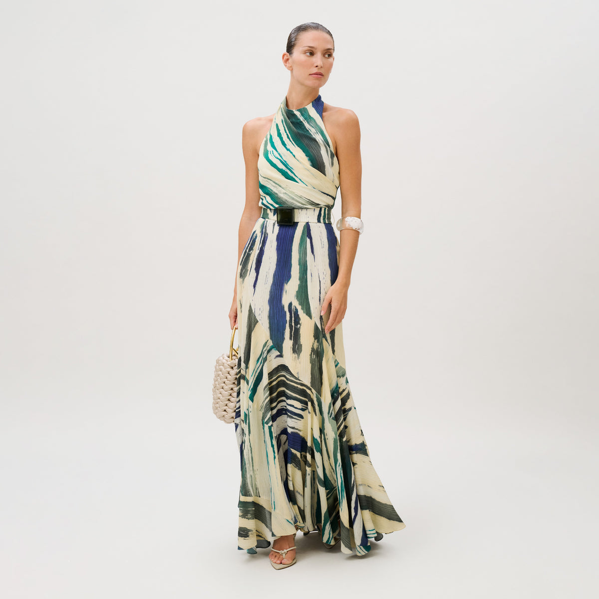 A model wears the Marwa Dress, a sleeveless, high-neck crepe de viscose maxi featuring a blue, green, and cream abstract print, cinched with a wide metallic belt and styled with a woven handbag.