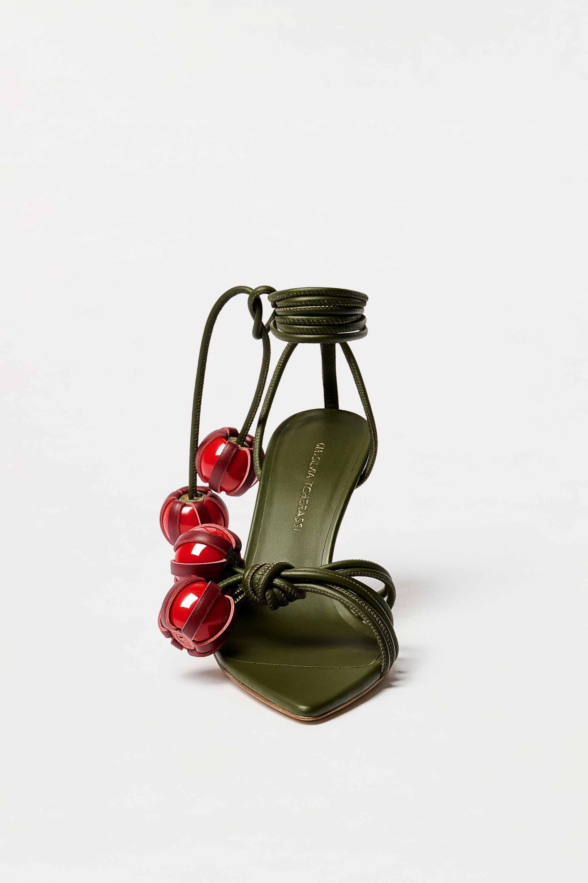The Mele Heels are olive green high-heeled sandals, expertly crafted from Italian leather, featuring ankle ties adorned with large red bead accents and displayed against a plain white background.
