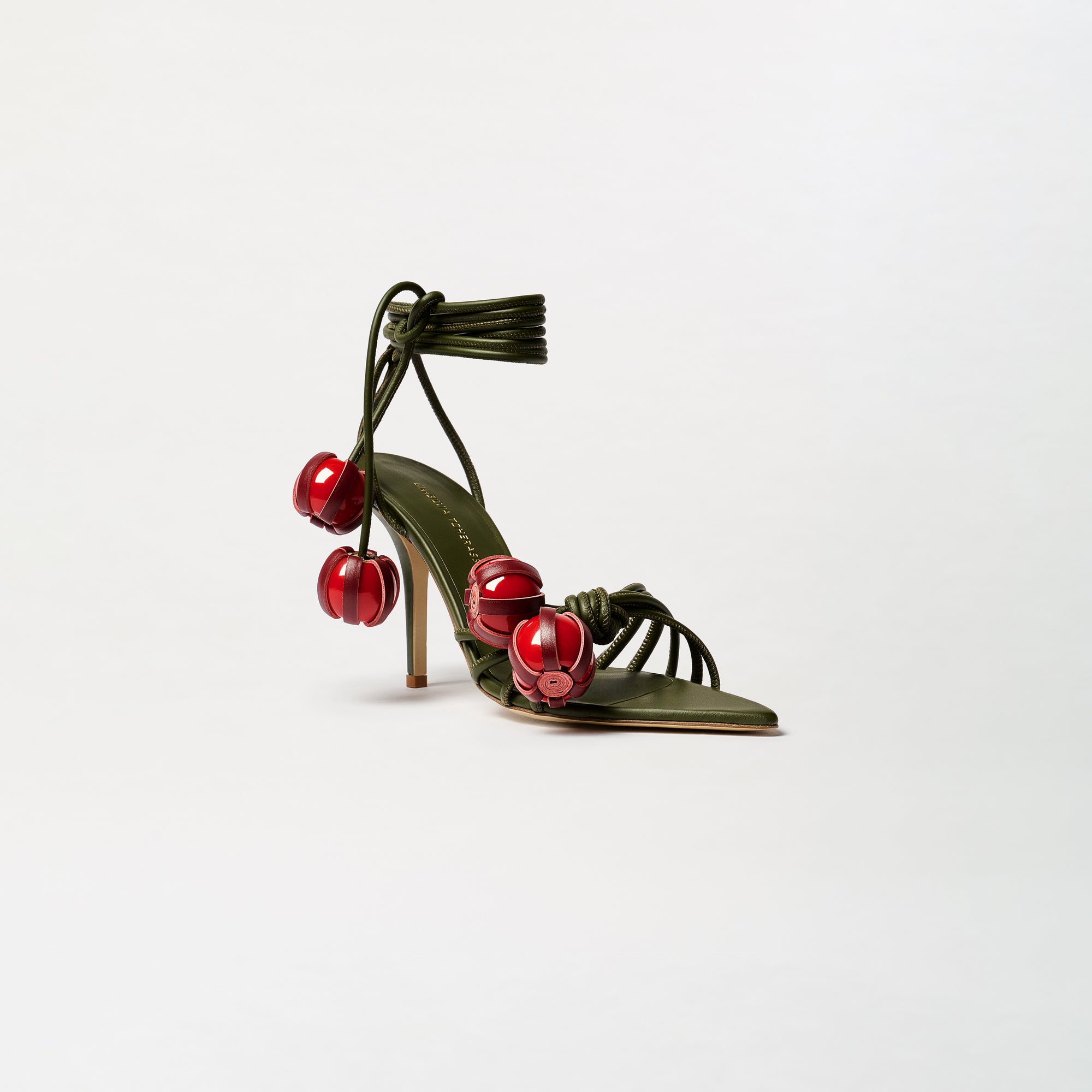 The Mele Heels are high-heeled sandals, expertly crafted from Italian leather, featuring olive green straps adorned with large red bead embellishments and an elegant ankle wrap tie, displayed on a plain white background.