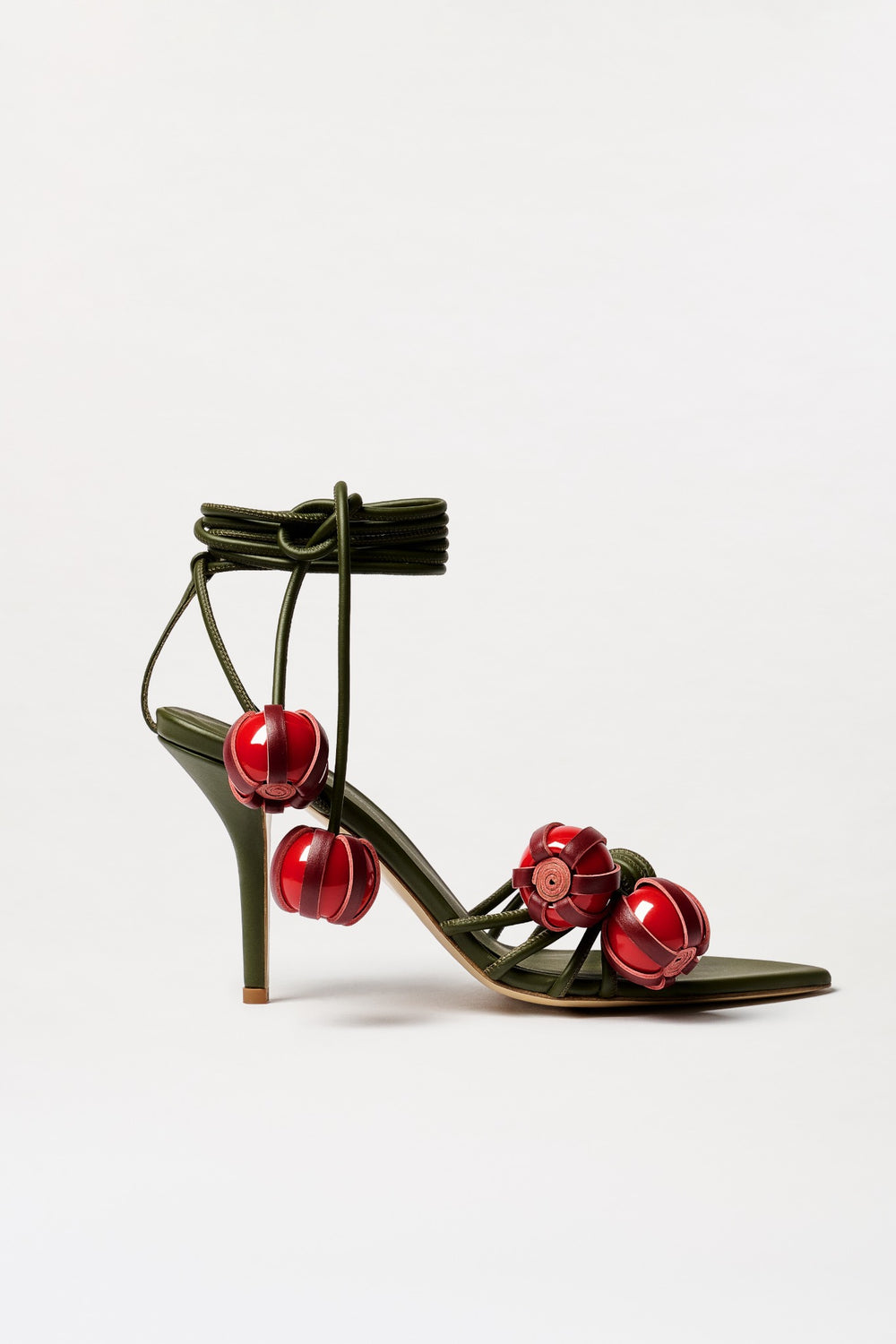 The Mele Heels are elegant olive green high-heeled sandals, expertly crafted in Italy from luxurious Italian leather. Featuring ankle wrap straps and three large red decorative ornaments adorning the front straps, these stunning heels are shown against a plain white background.