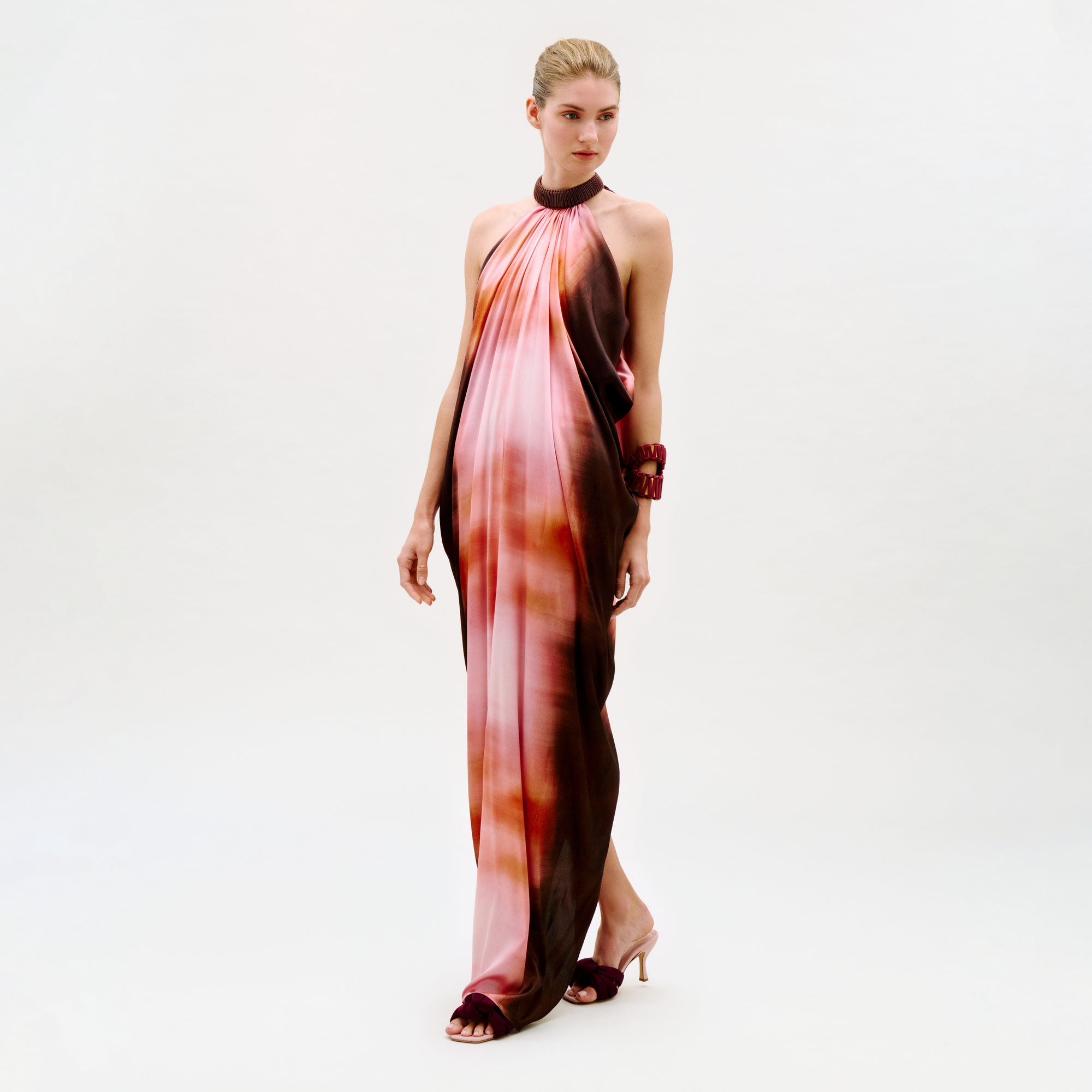 A woman stands against a plain light background wearing the Mene Dress, a flowing halter-neck gown in pink, orange, and brown tones. Her hair is pulled back, and she accessorizes with a maroon bracelet and matching heeled sandals.