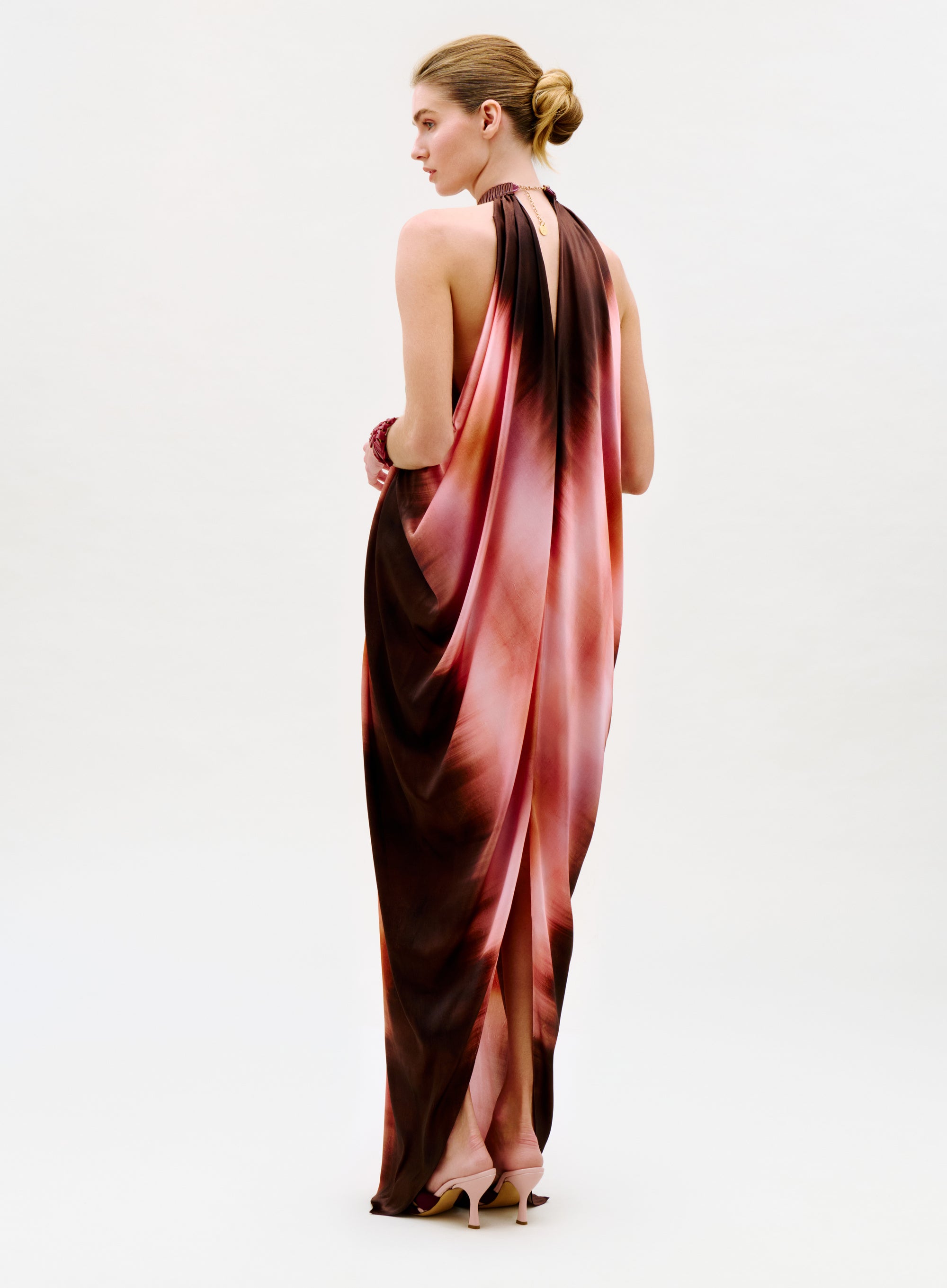 A woman with blonde hair in a low bun wears the Mene Dress—a flowing, sleeveless pink and brown halter-neck gown—standing sideways and glancing over her shoulder against a plain light background.