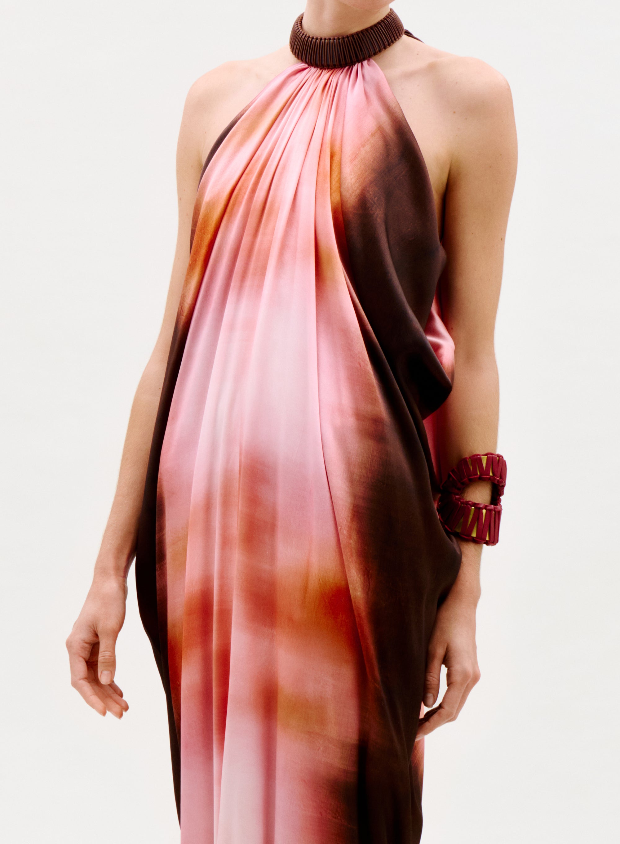Someone wearing the Mene Dress, a flowing halter-neck piece with a pink and brown gradient, styled with a chunky dark choker and matching statement bracelet, set against a plain white background.
