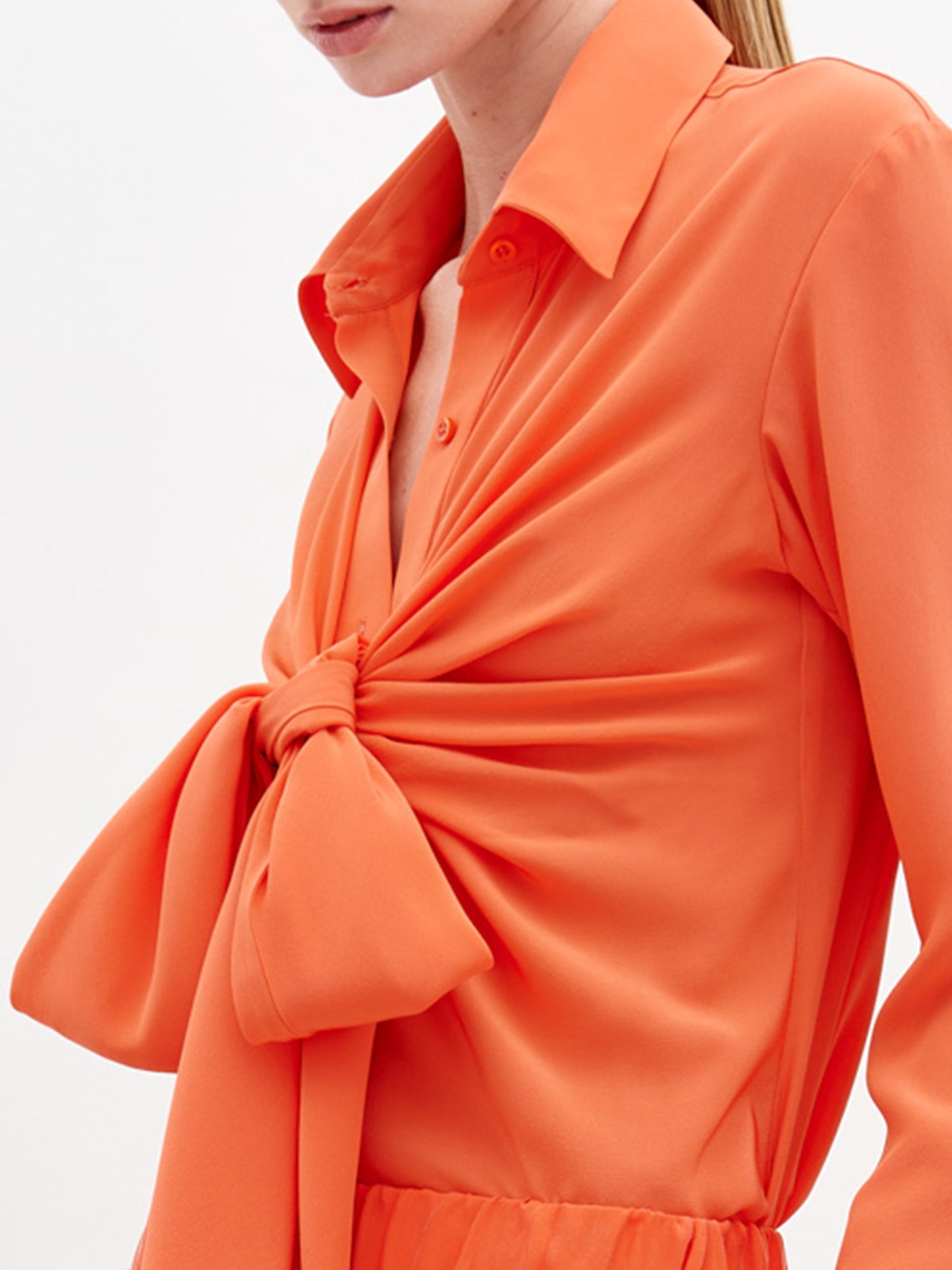 A woman wears the Mercury Blouse Tangerine in orange silk, crafted from fine Italian textile with a large front knot revealing part of her chest. The image is cropped to show only her torso and lower face.