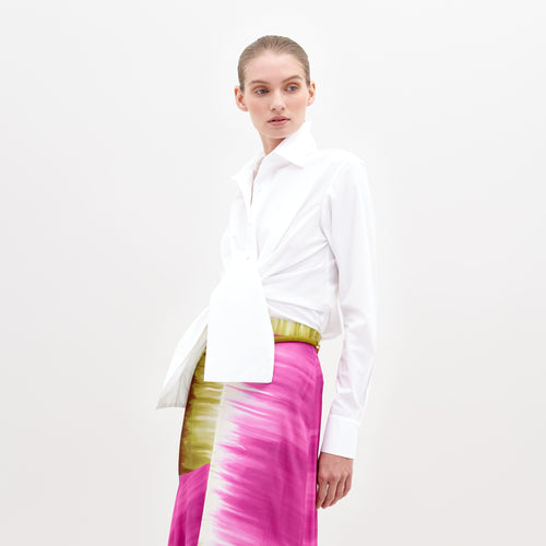 A person wearing a Mercury Blouse White and a vibrant pink skirt with green accents, standing against a plain white background, available for pre-order now. Ships by November 15th, 2024.