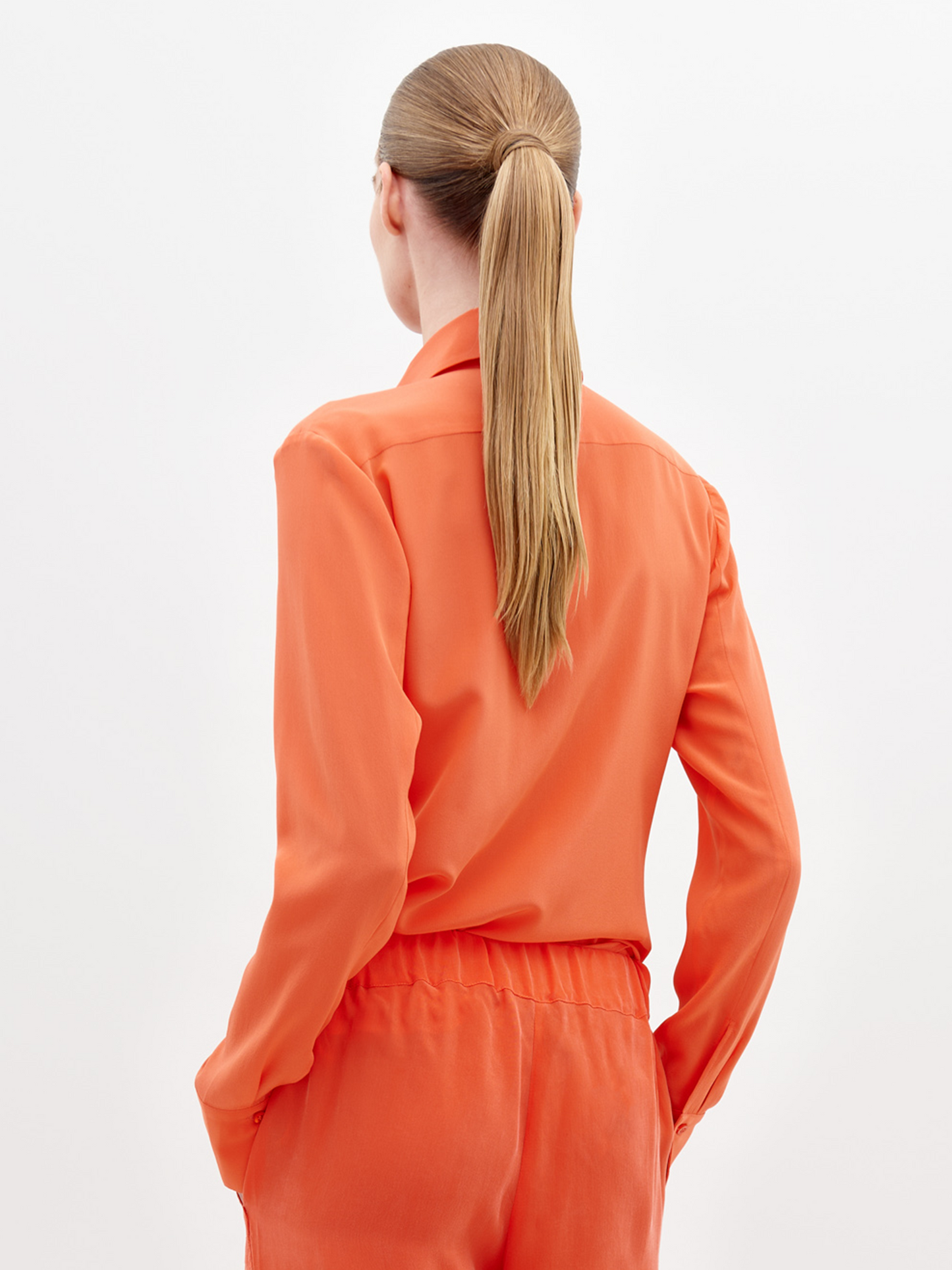 A person with long, straight blonde hair in a ponytail wears the Mercury Blouse Tangerine, seen from behind against a plain white background.