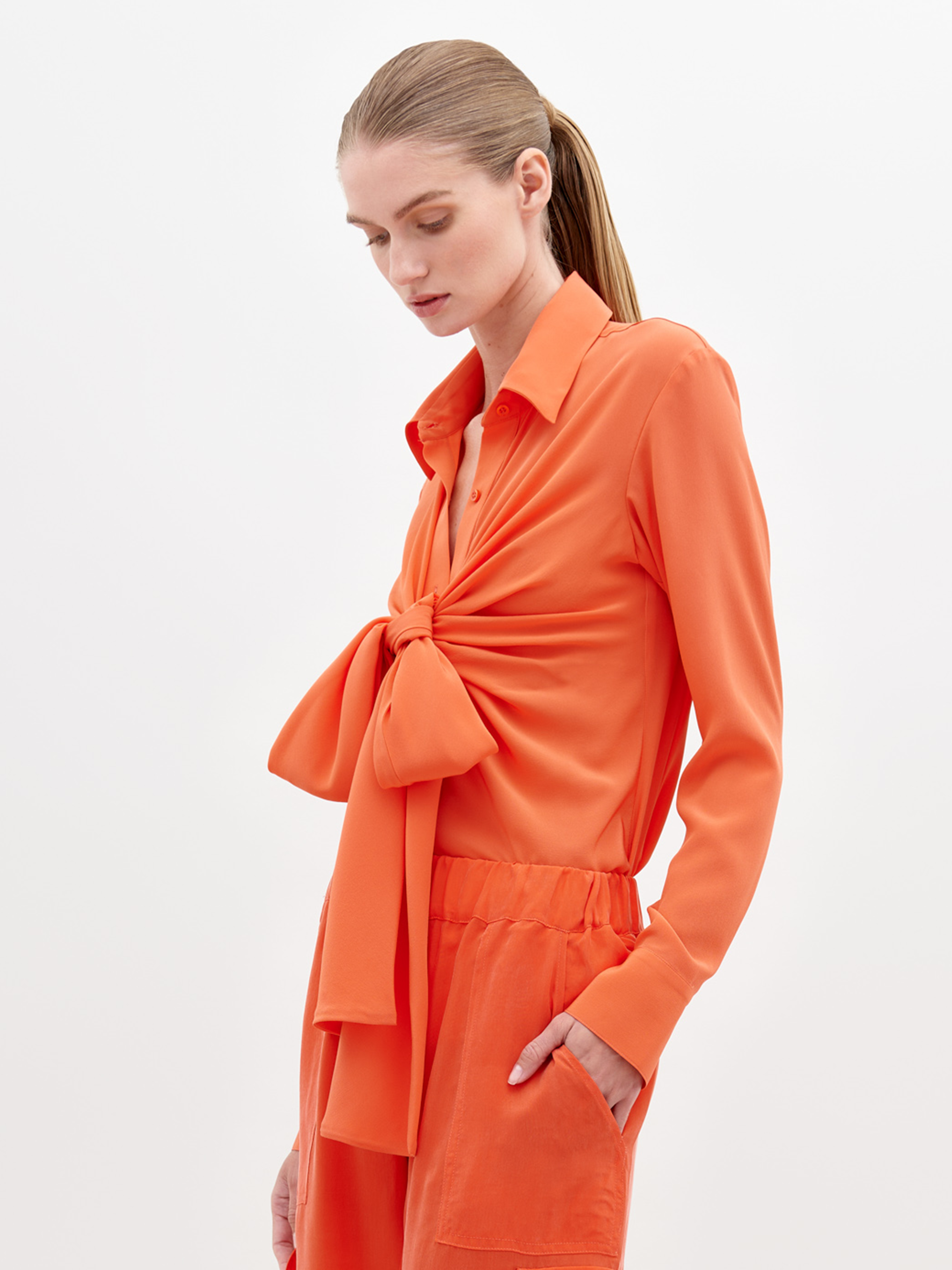 A woman with straight, light brown hair in a ponytail wears the Mercury Blouse Tangerine, styled with matching orange pants. She looks down neutrally against a plain white background.