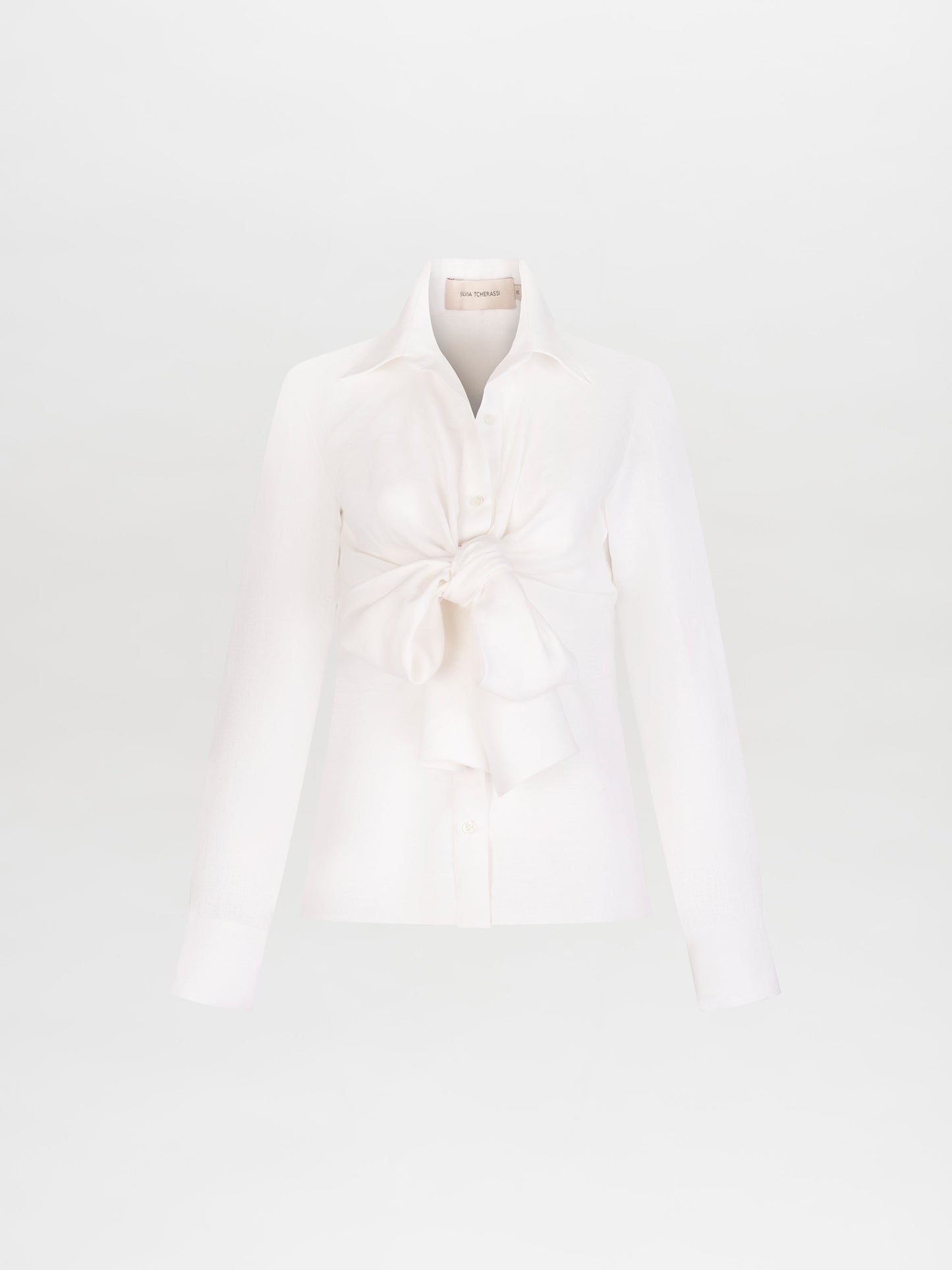 A person with long light brown hair in a ponytail is wearing the Mercury Blouse (Linen) White with a large bow, white pants, and holding beige shoes. Pre-order now to ensure you get this look by the 2025 ship-by date.