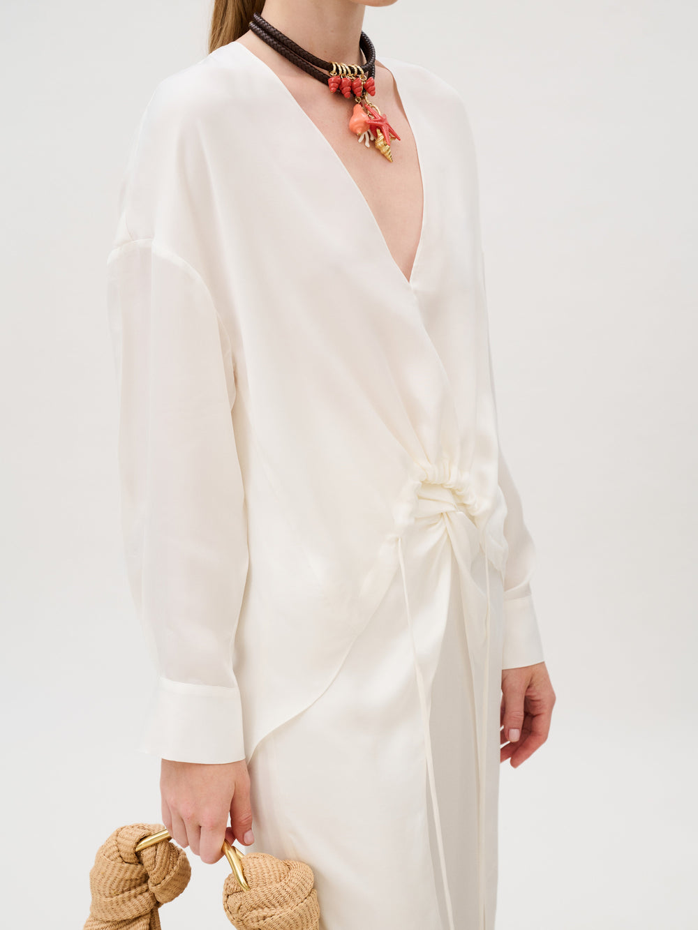 The Merida Blouse is a long-sleeved white Cupro top with a deep V-neck and cinched front detail, styled with a layered beaded necklace featuring coral accents and a woven straw handbag with gold handles.