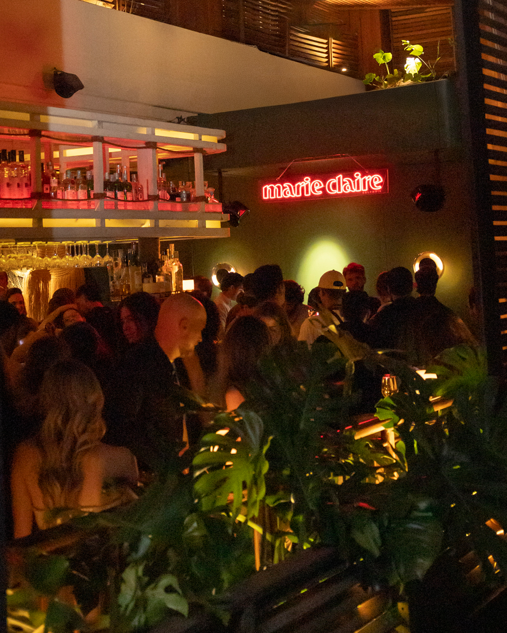A crowded indoor bar scene with people gathered around a counter and a glowing red neon sign reading 'marie claire' on the wall. Green plants and warm ambient lighting are visible in the foreground and background.