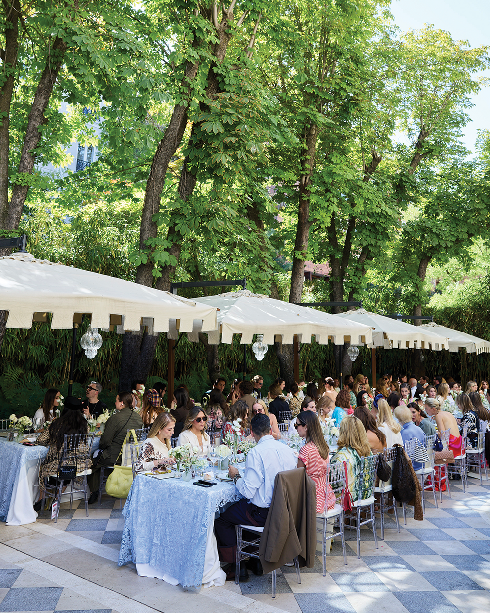 Outdoor event with people seated at tables under large umbrellas in a park-like setting.
