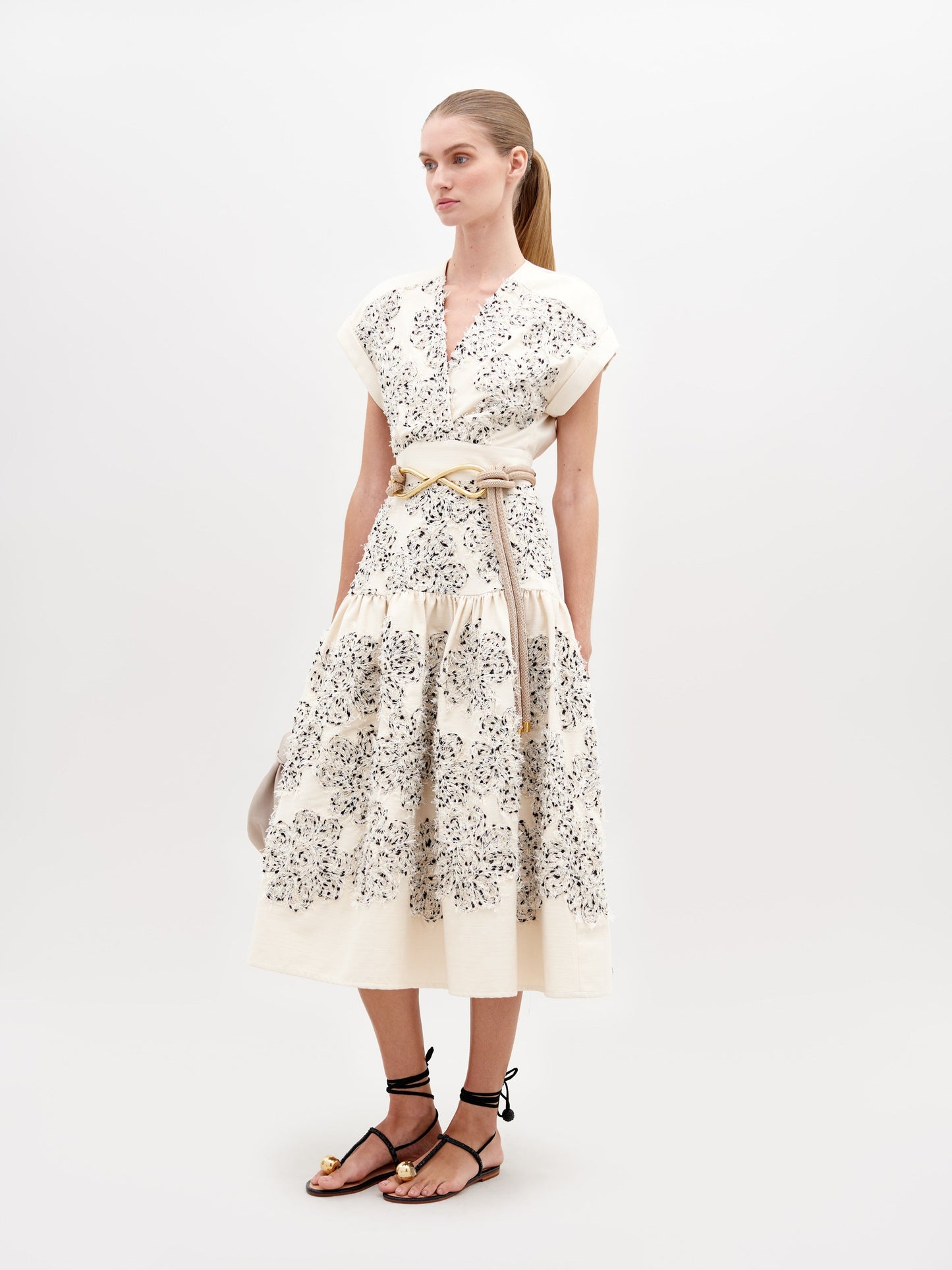 A woman wearing a Metaponto Dress Beige Black Embroidered Flowers with a beige belt and black sandals stands against a plain white background, ready for the fashion pre-order set to ship by date November 15th, 2024.