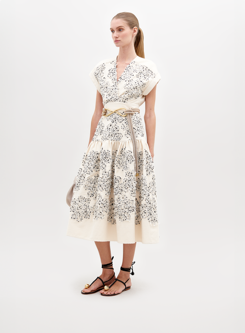 A woman wearing a Metaponto Dress Beige Black Embroidered Flowers with a beige belt and black sandals stands against a plain white background, ready for the fashion pre-order set to ship by date November 15th, 2024.