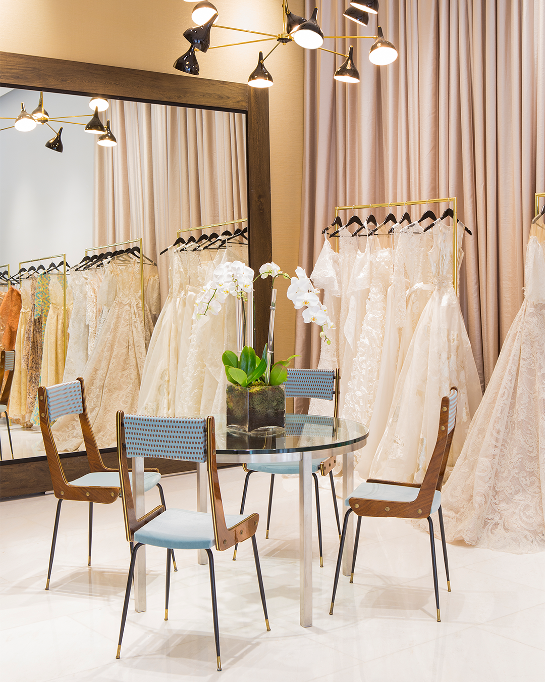 A bridal boutique with a glass table and chairs in front of a mirror. Several wedding dresses hang on racks with beige curtains in the background.