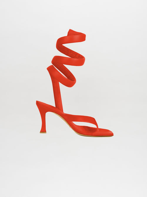 The Mika Heels Red feature a striking red hue, crafted from luxurious Italian calf leather with an elegant spiral ankle strap, set against a minimalistic background.