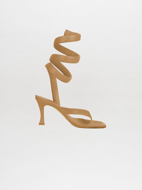 The Mika Heels Taupe are designed with a spiral ankle strap and crafted from premium Italian calf leather in an elegant taupe color, contrasted beautifully against a clean white background.