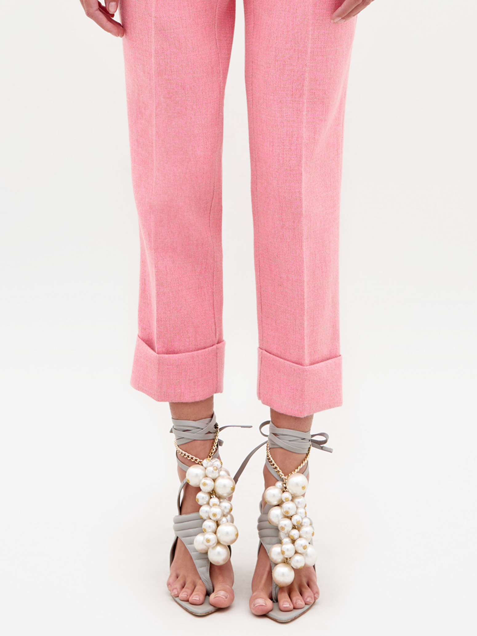 A person stands against a plain background, wearing the Moad Pant Pink suit, featuring a tailored cropped jacket and matching pink pants. They complete their look with high-heeled shoes adorned with white embellishments, ready for shipment by February 15th, 2025.