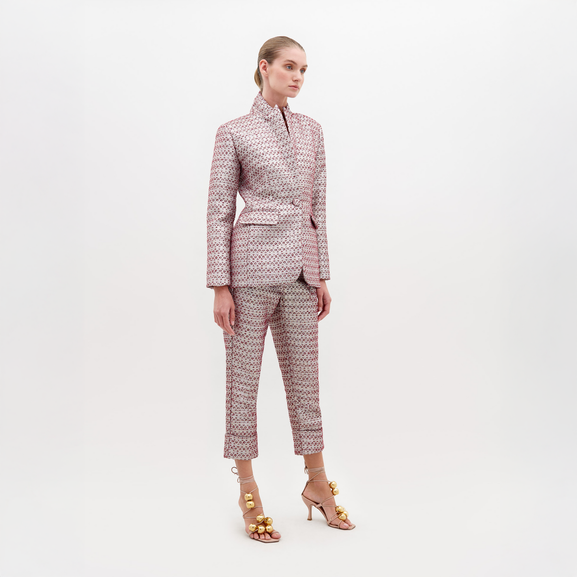 A woman stands against a plain background, wearing a patterned suit with the Moad Pant Burgundy and a tied belt. She is also wearing high-heeled sandals adorned with gold baubles. Pre-order by November 15th, 2024, to ensure it ships by date.