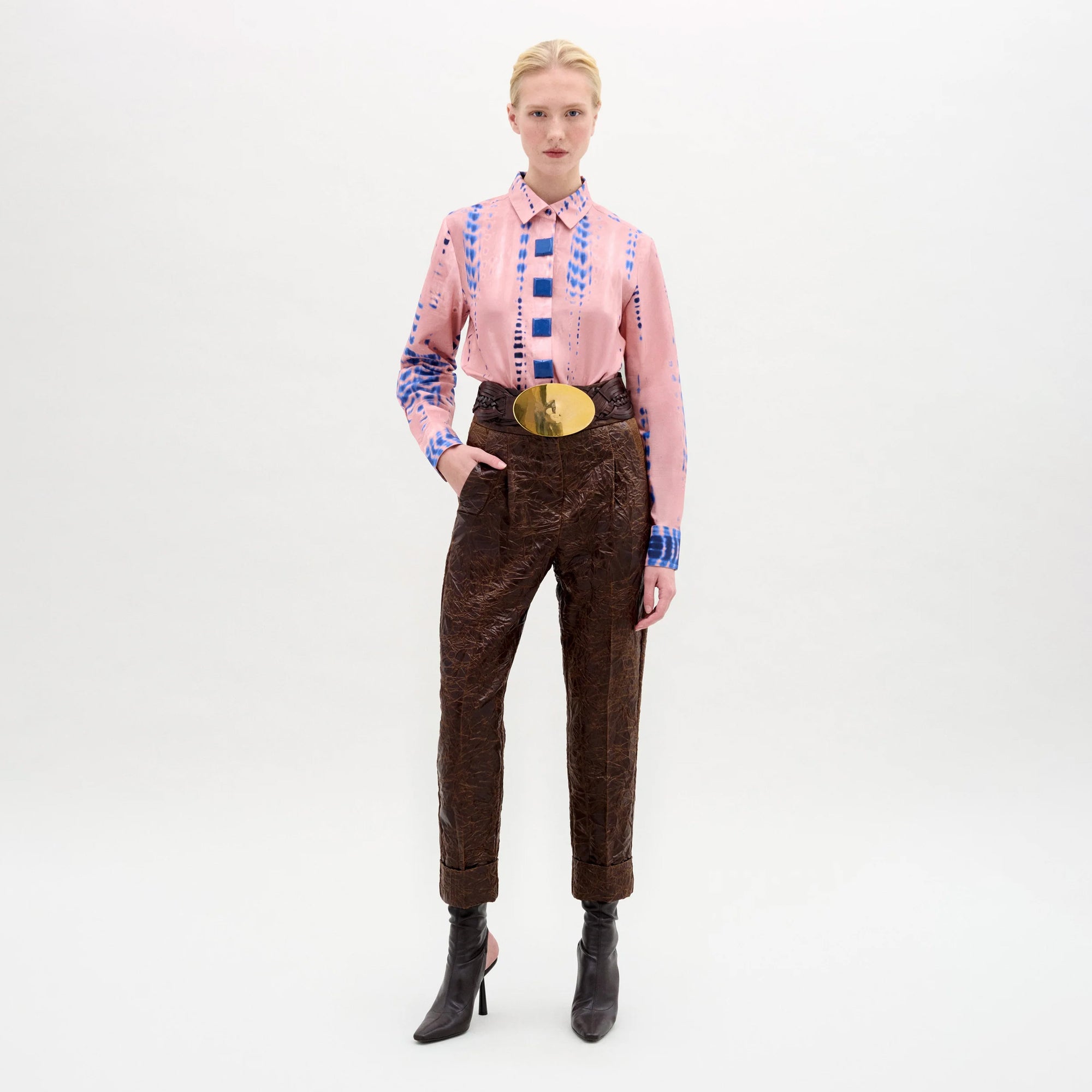 A person stands against a plain white background wearing the Moad Pant, a high-waisted, straight-leg, cropped style in Italian fabric, styled with a pink patterned shirt, large gold belt, and pointed dark ankle boots. One hand is in their pocket.